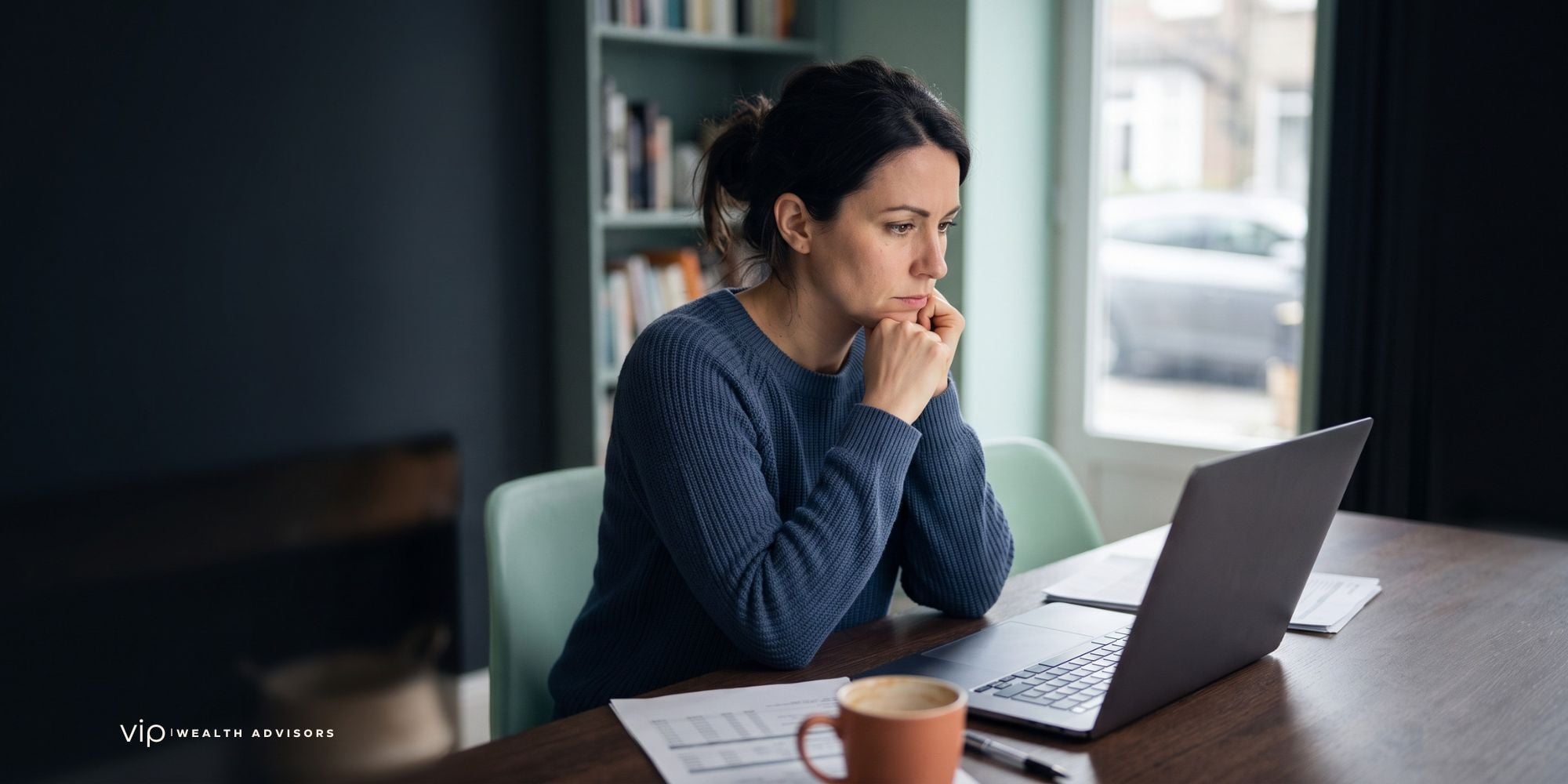 A thoughtful woman sits at a desk in a modern office, looking intently at her laptop screen with financial documents and a coffee mug in front of her.